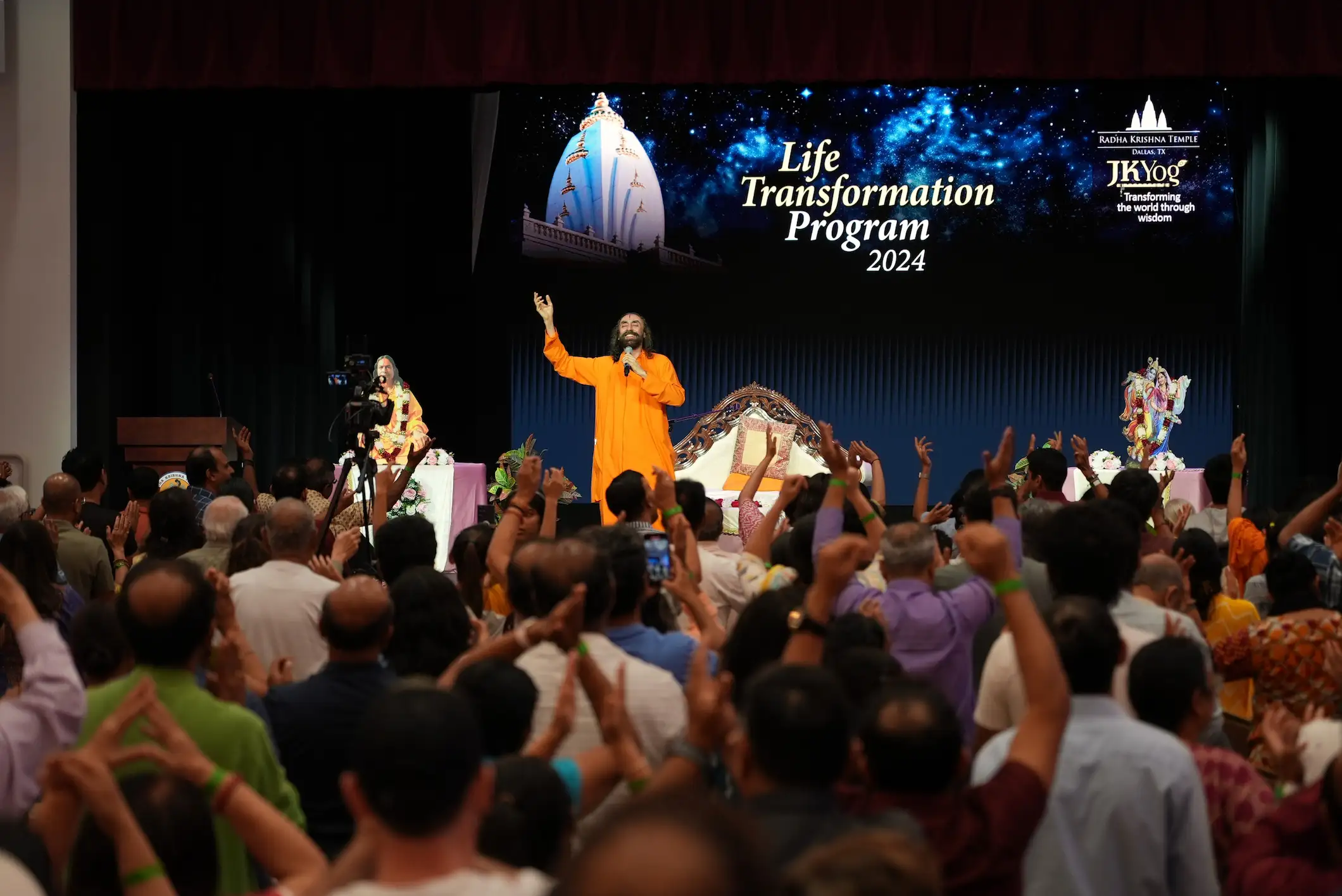 Swami Mukundananda addressing a large audience during the Life Transformation Program 2024, with attendees engaged in spiritual practice and reflection.