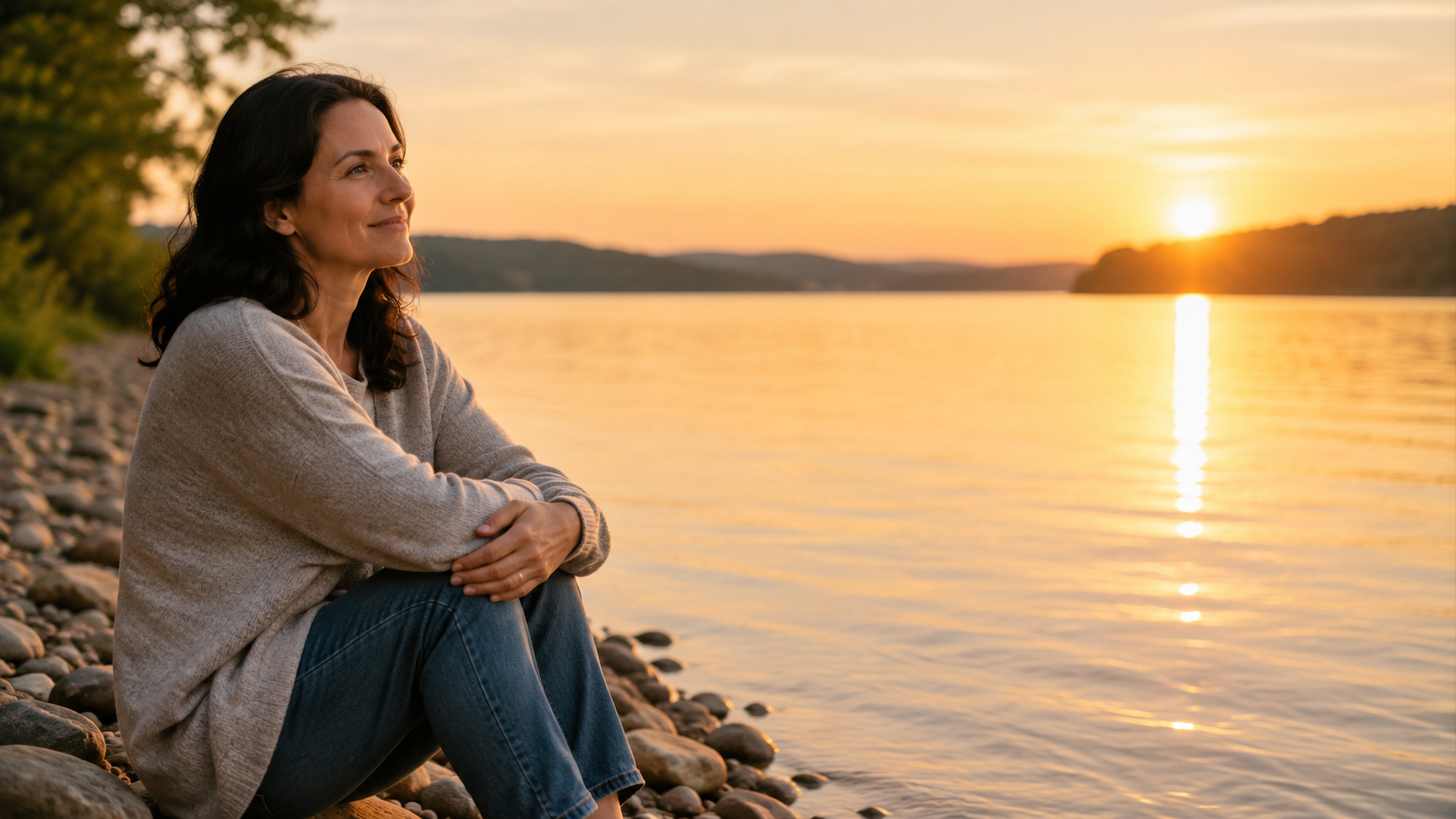 A woman sits peacefully on a rocky lakeside at sunset, looking toward the glowing water and horizon with a calm, reflective expression.