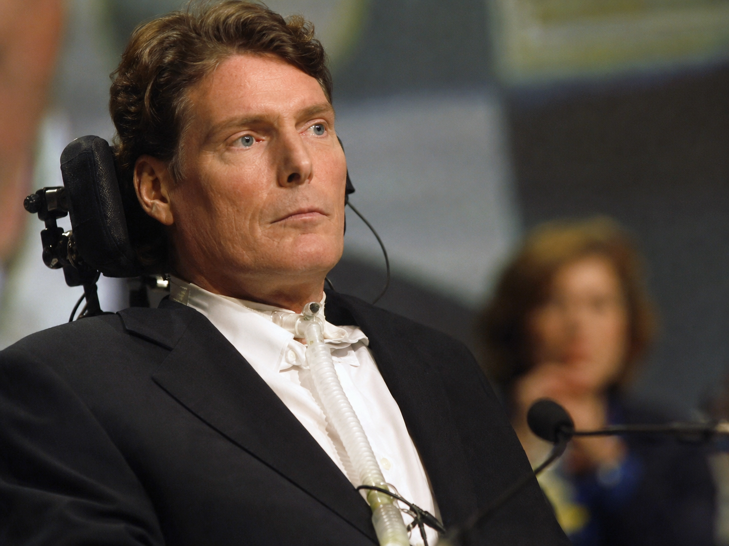 A realistic image of Christopher Reeve seated in a wheelchair at a formal public event, wearing a dark suit with a ventilator tube visible, representing his life after the 1995 spinal cord injury.