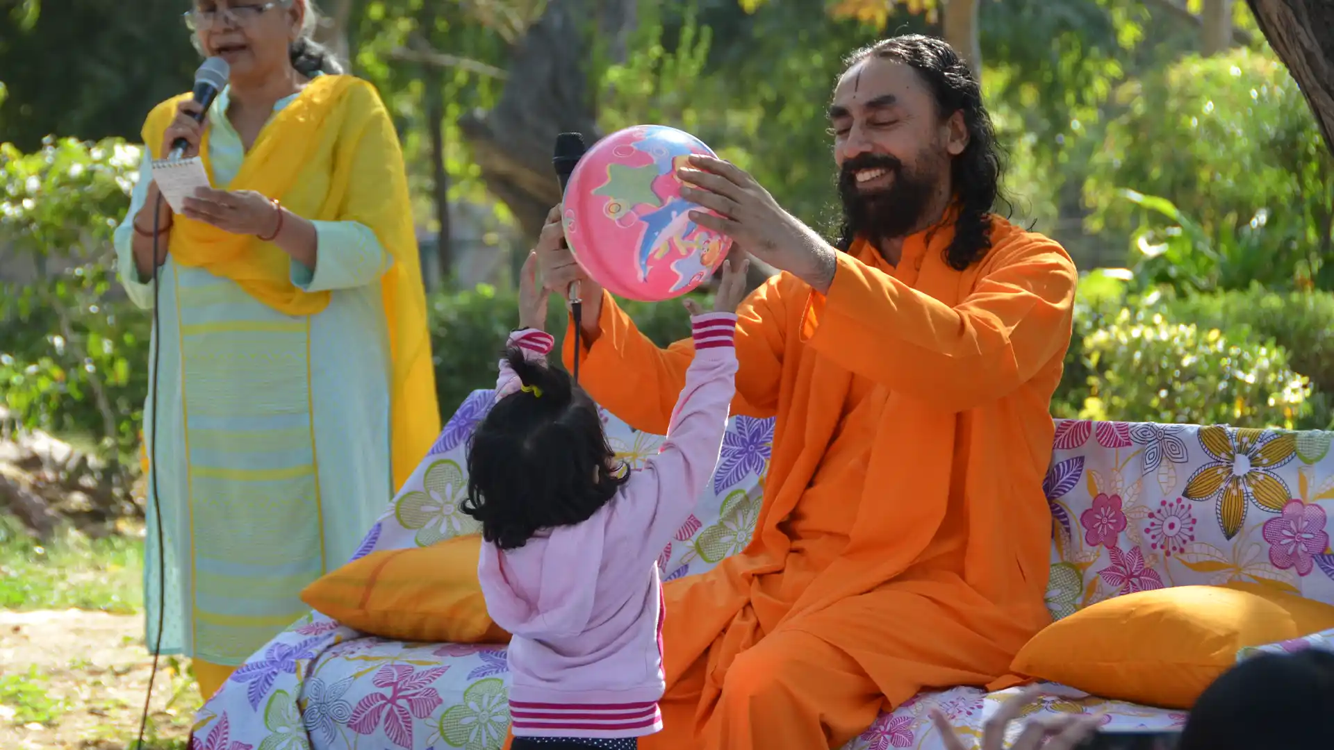 Swami Mukundananda sits outdoors in a peaceful green setting, smiling as he warmly interacts with a young child holding a colorful balloon, symbolizing mentorship, guidance, joy, and the supportive wisdom that helps human growth.