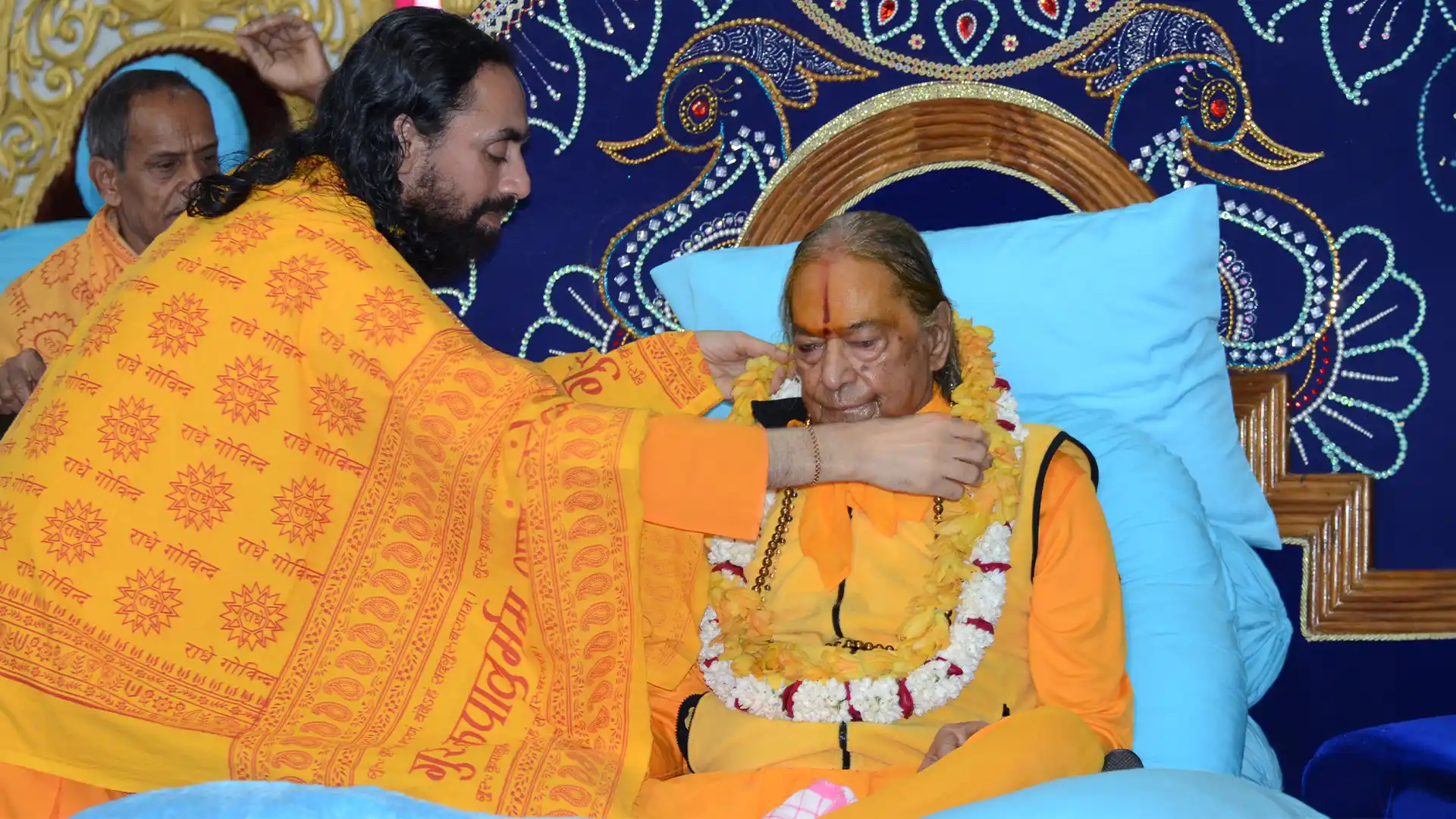Swami Mukundananda lovingly places a garland around his Guru, Jagadguru Kripaluji Maharaj, in a devotional setting, symbolizing reverence, humility, surrender, and the spiritual necessity of a Guru as the remover of inner darkness.