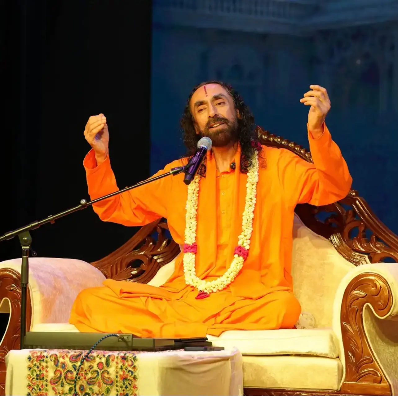 Spiritual discourse at the Dallas Spiritual Retreat, with a saffron-robed Swami Mukundananda speaking into a microphone while seated on stage.